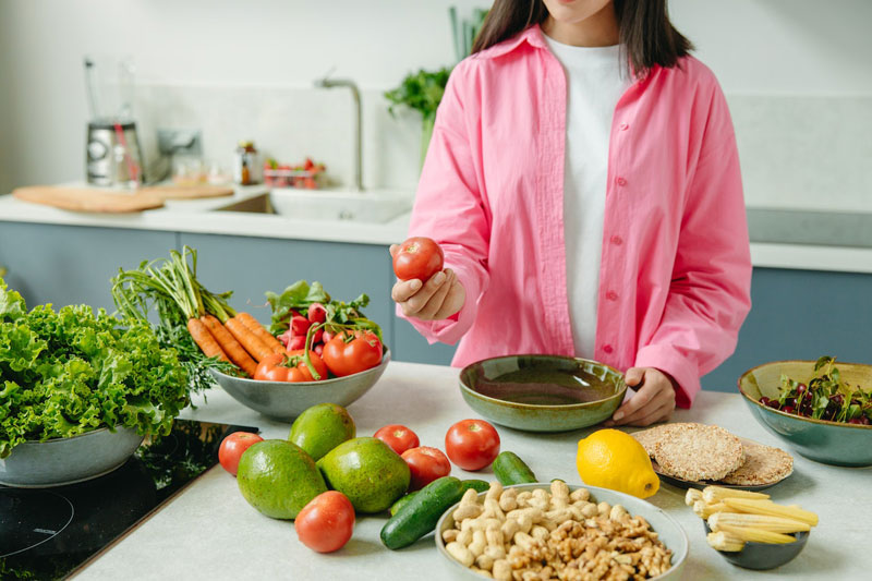 A woman holding a tomato in the kitchen with lots of different vegetables on the counter.